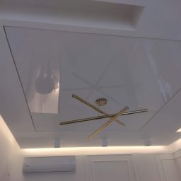Usługi Remontowo Budowlane Kamil Krawczyk - Biały, błyszczący sufit z wbudowanym oświetleniem LED i nowoczesną, złotą lampą wiszącą z rurek.