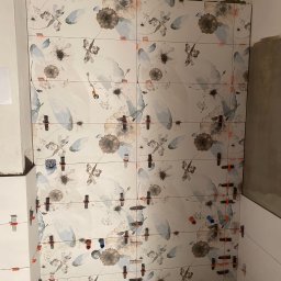 Usługi Remontowo Budowlane Kamil Krawczyk - Ściana w trakcie układania płytek z motywem kwiatowym, z użyciem poziomych sznurków i klipsów dystansowych, widoczne niedociągnięcia przy suficie i obok ściany z białych płytek.