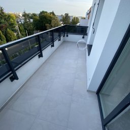 Usługi Remontowo Budowlane Kamil Krawczyk - Jasne płytki na balkonie z czarną balustradą, widok na zieleń drzew i budynki w oddali, białe ściany budynku. Kabel elektryczny zwisający ze ściany.