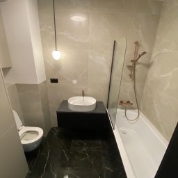 Usługi Remontowo Budowlane Kamil Krawczyk - Nowoczesna łazienka z czarną marmurową podłogą, beżowymi płytkami imitującymi marmur na ścianach, białą umywalką nablatową na czarnej szafce, toaletą i białą wanną z parawanem oraz armaturą...