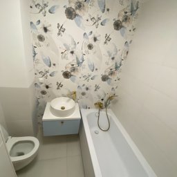 Usługi Remontowo Budowlane Kamil Krawczyk - Wykończona łazienka z białą ceramiką, umywalką nablatową na błękitnej szafce, wanną i toaletą, akcentowana tapetą w kwiatowe wzory i złotą armaturą.