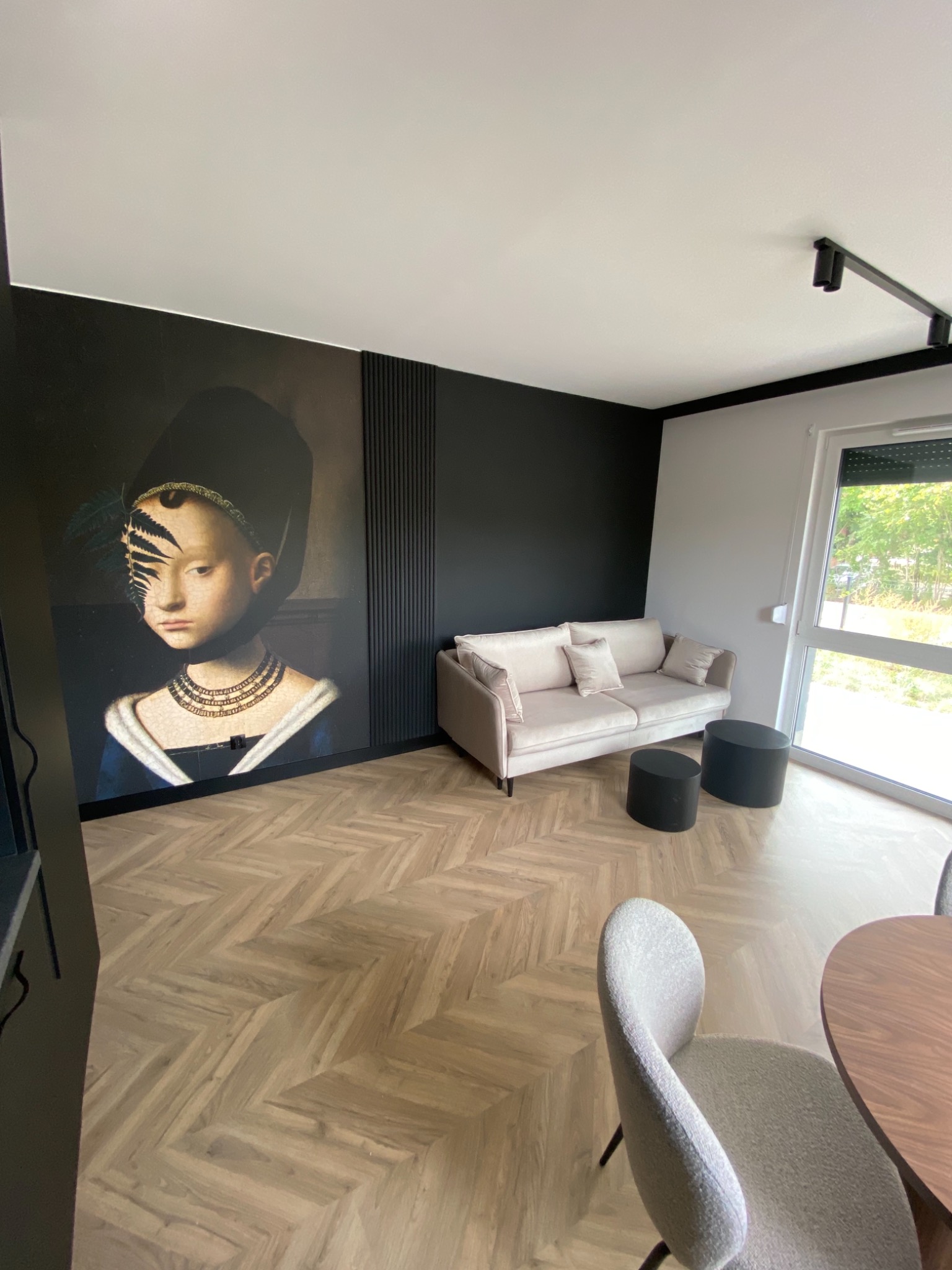 Salon z panelami ułożonymi w jodełkę, czarną ścianą z dekoracyjnymi pionowymi listwami i fototapetą przedstawiającą portret kobiety z paprocią na twarzy, beżową sofą i okrągłymi stolikami kawowymi.