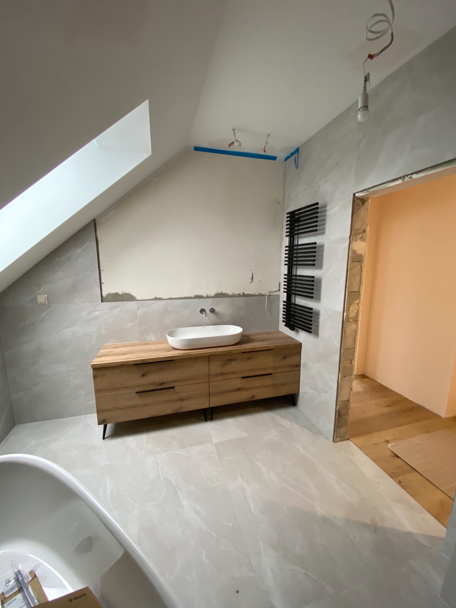 Łazienka na poddaszu w trakcie wykańczania: drewniana szafka pod umywalką, grzejnik drabinkowy, ściany wyłożone płytkami imitującymi marmur, widoczne instalacje i surowy stan nad szafką.