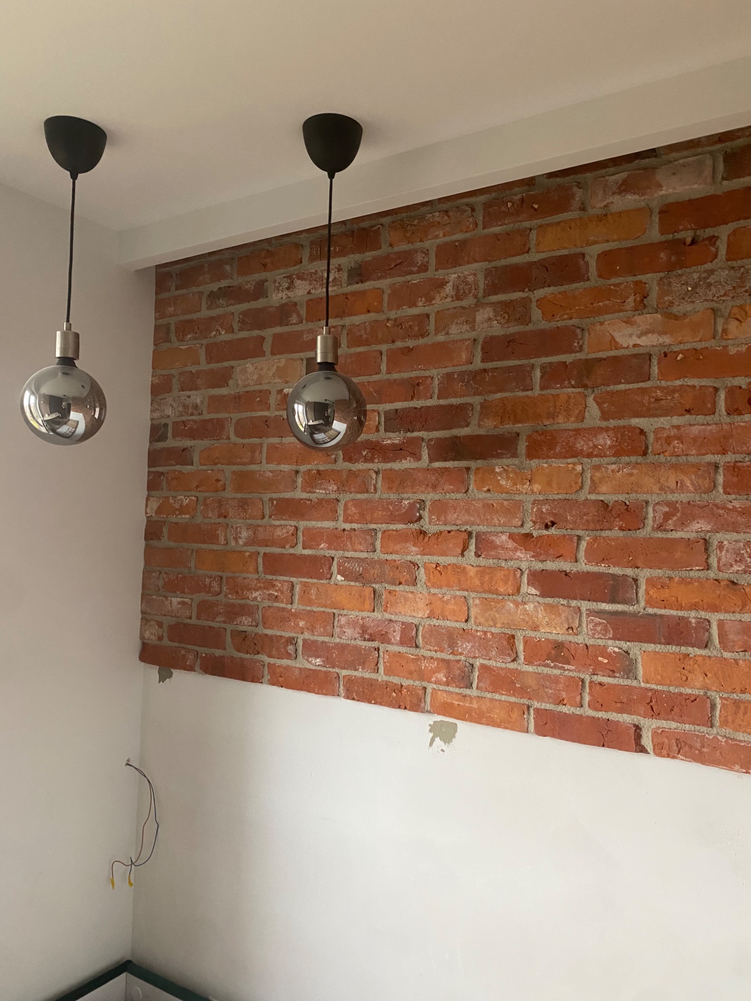 Częściowo wykończona ściana z czerwonej cegły w pomieszczeniu z dwoma srebrnymi lampami wiszącymi, widoczne przewody elektryczne i niedoskonałości w malowaniu.