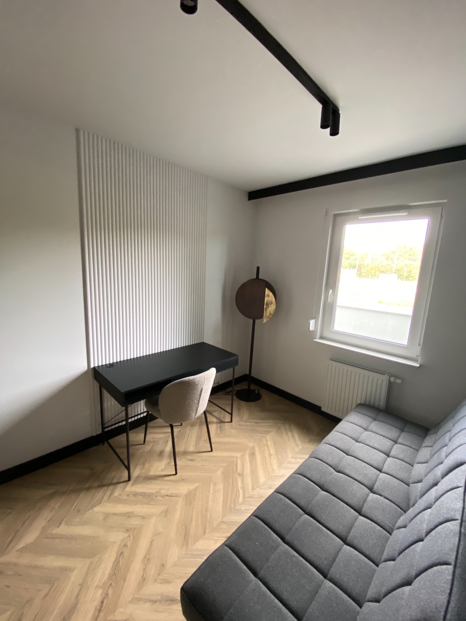 Nowoczesne wnętrze pokoju z panelem dekoracyjnym na ścianie, biurkiem z krzesłem, szarą sofą i parkietem ułożonym w jodełkę, oświetlone lampą sufitową.