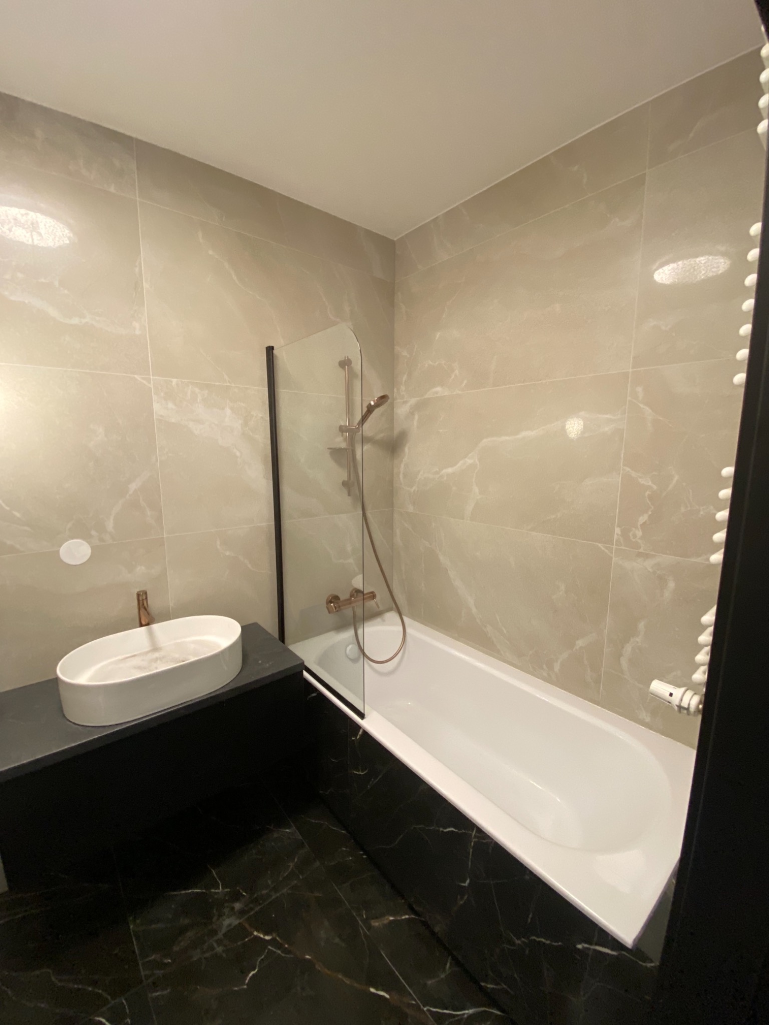 Wykończona łazienka z beżowymi płytkami imitującymi marmur na ścianach, czarną marmurową podłogą, owalną umywalką na czarnym blacie i białą wanną z parawanem.