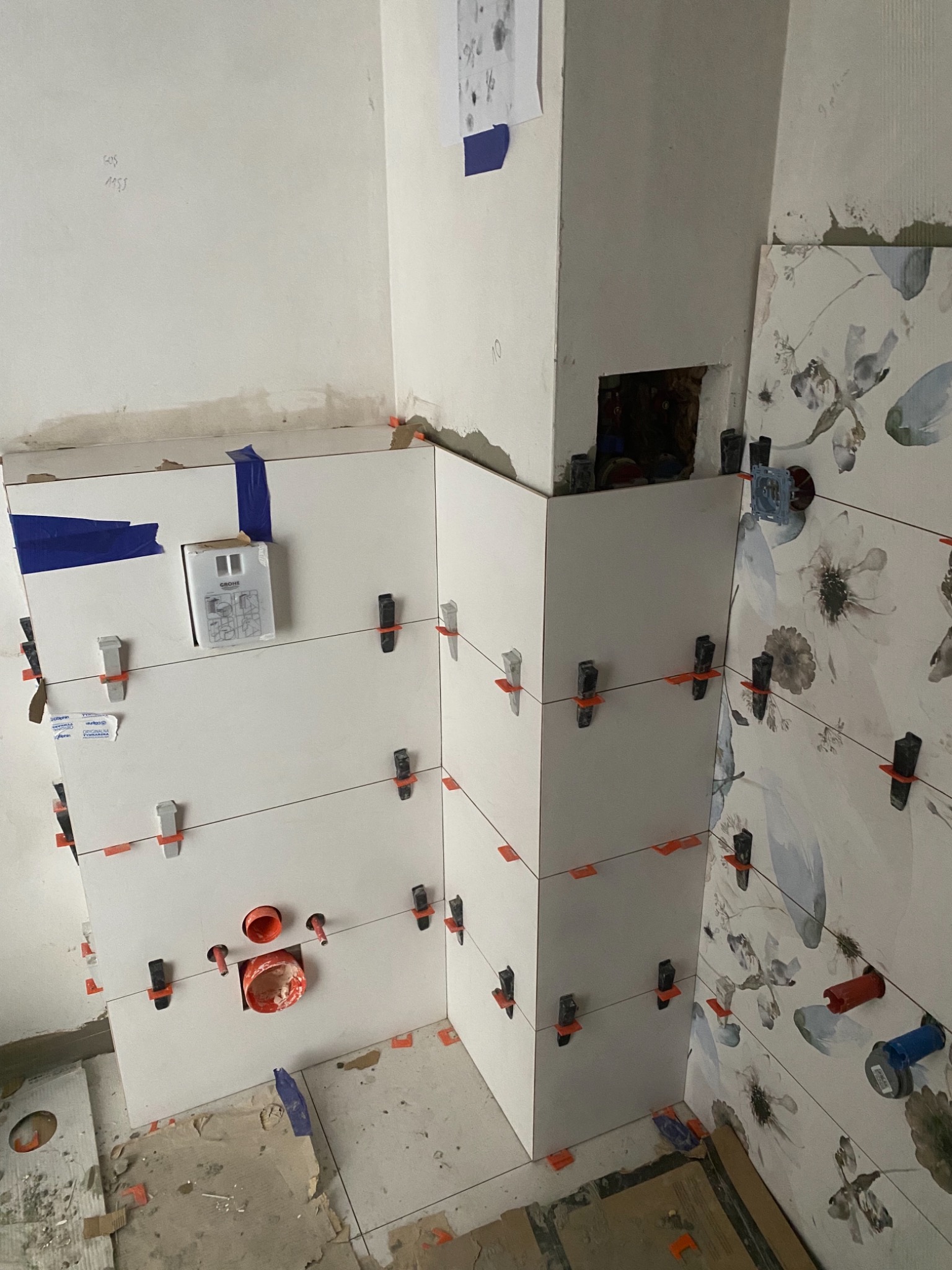 Montaż białych i wzorzystych płytek w narożniku łazienki, z użyciem klipsów poziomujących i taśmy maskującej, widoczne elementy instalacji wodnej i fragmenty podłoża.