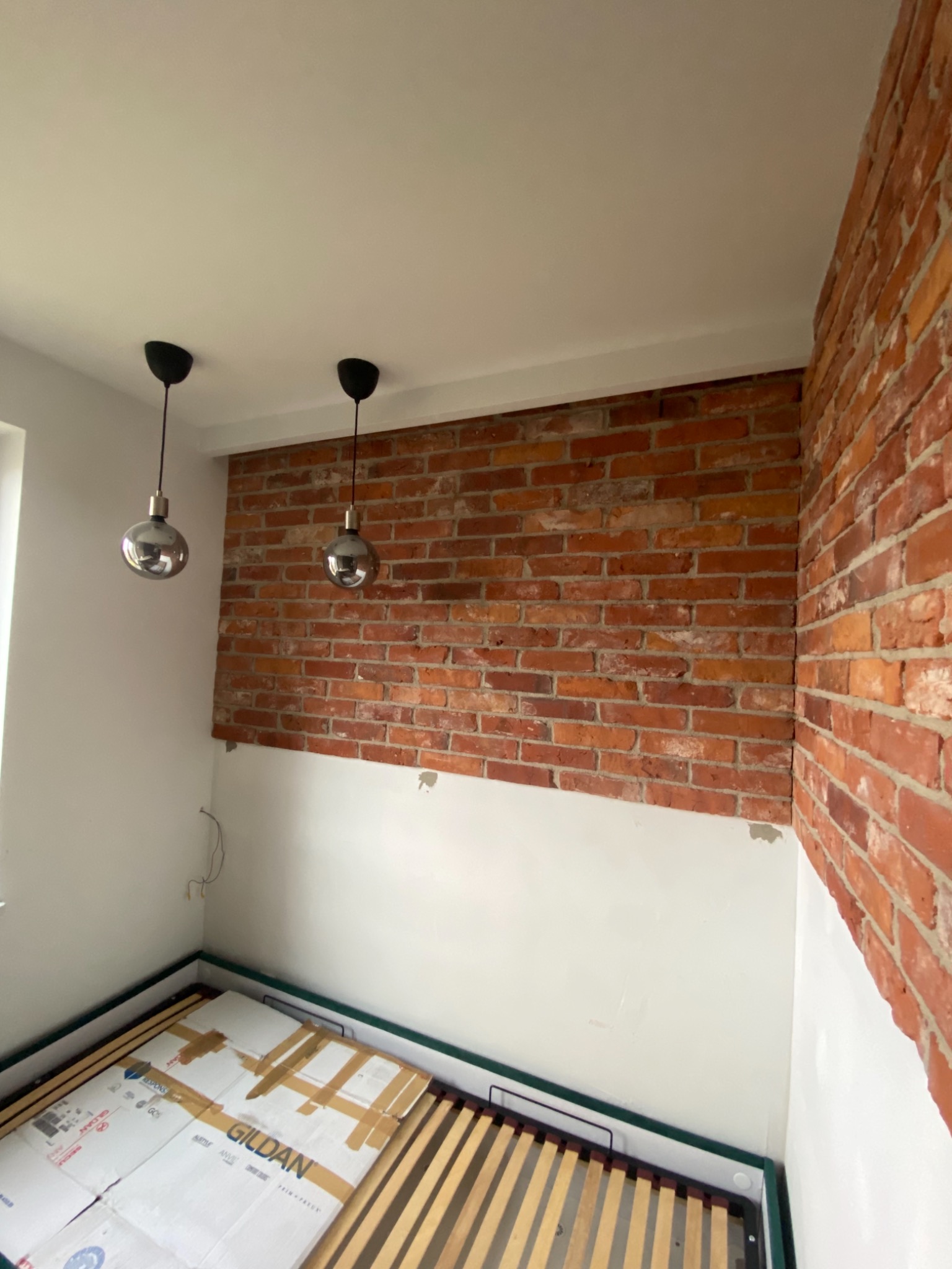 Ściana z czerwonej cegły w trakcie remontu pokoju, widoczne niedomalowane fragmenty ściany i sufitu, oraz fragment łóżka z kartonami