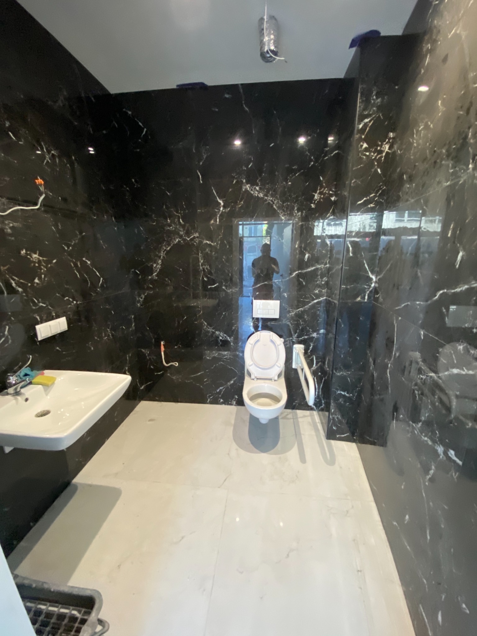 Nowoczesna łazienka w trakcie wykańczania z czarnymi płytkami imitującymi marmur na ścianach i jasnymi płytkami na podłodze. Widoczna umywalka, toaleta z podwieszanym systemem spłukiwania...