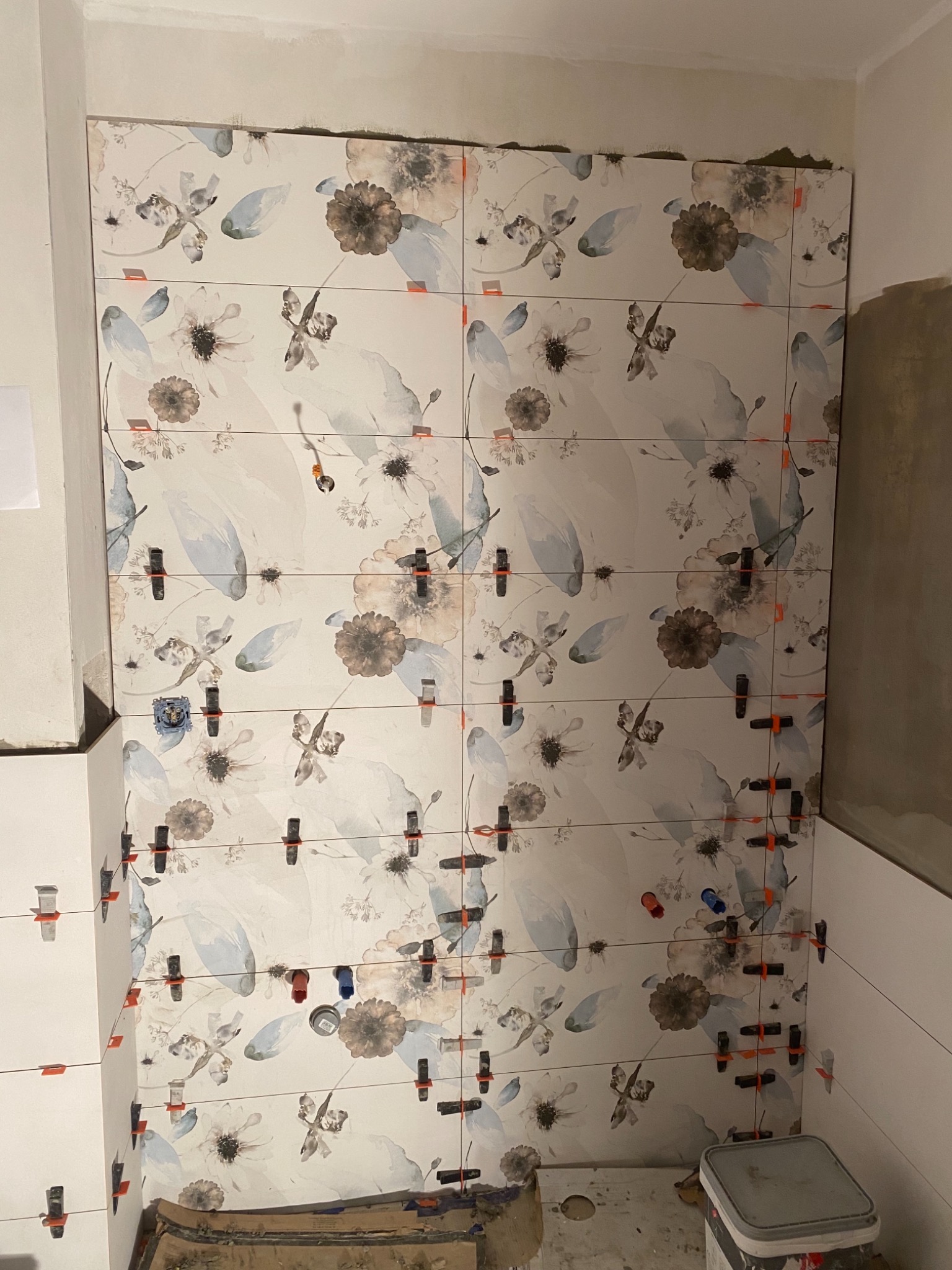 Ściana w trakcie układania płytek z motywem kwiatowym, z użyciem poziomych sznurków i klipsów dystansowych, widoczne niedociągnięcia przy suficie i obok ściany z białych płytek.