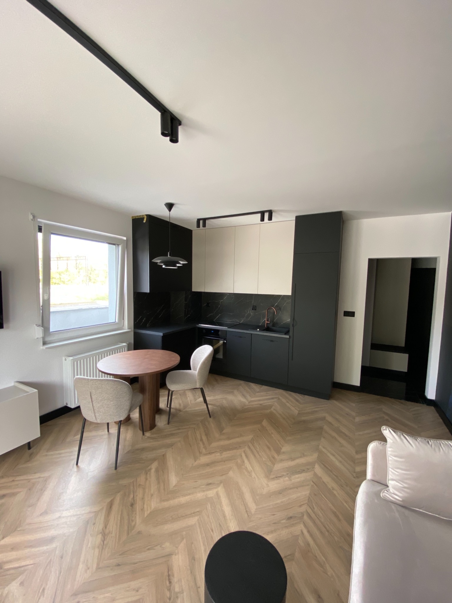 Nowoczesna aranżacja wnętrza w Toruniu: ciemna zabudowa kuchenna z marmurowym blatem, drewniana podłoga ułożona w jodełkę, okrągły stół i krzesła w jasnych odcieniach, oświetlenie szynowe.