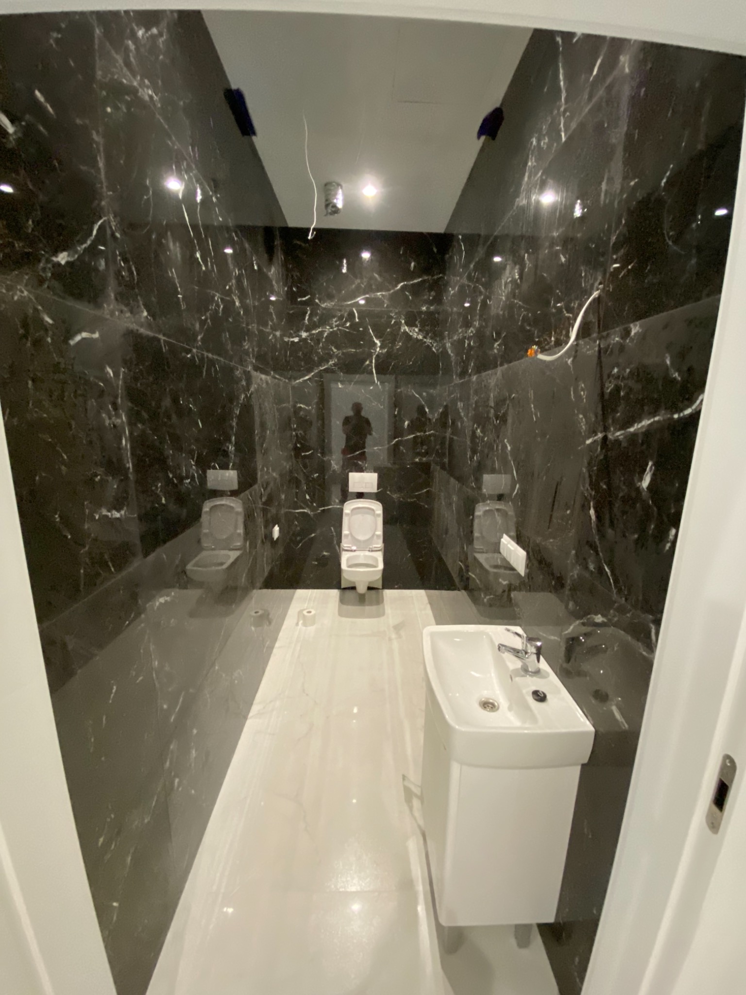 Wąska łazienka wykończona czarnym marmurem z białymi żyłami, trzy toalety wzdłuż ściany, biała umywalka z szafką, widoczne niedokończone prace elektryczne.