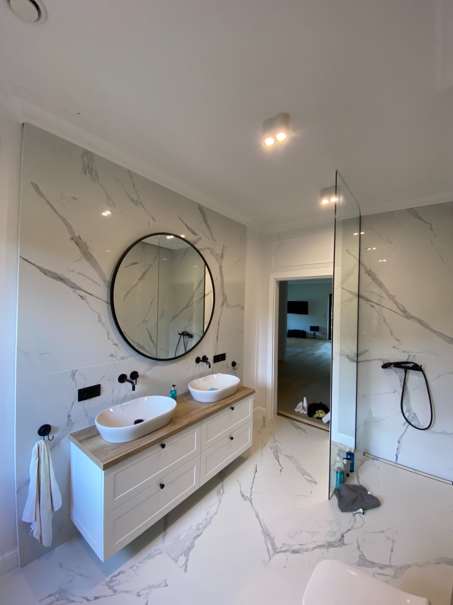 Nowoczesna łazienka z marmurowymi płytkami, owalnymi umywalkami nablatowymi i czarną armaturą, uzupełniona okrągłym lustrem i kabiną prysznicową typu walk-in.