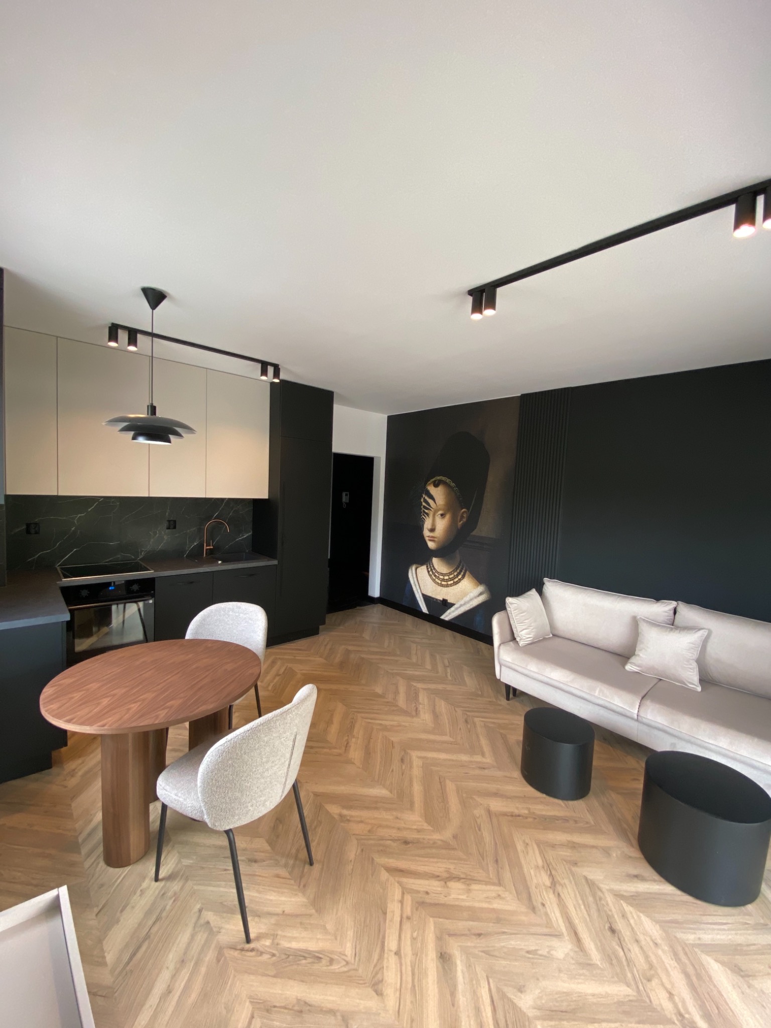 Jasne panele ułożone w jodełkę francuską, owalny drewniany stół z krzesłami, aneks kuchenny z marmurowym blatem i czarnymi szafkami, sofa z poduszkami i czarne stoliki kawowe na tle ściany...