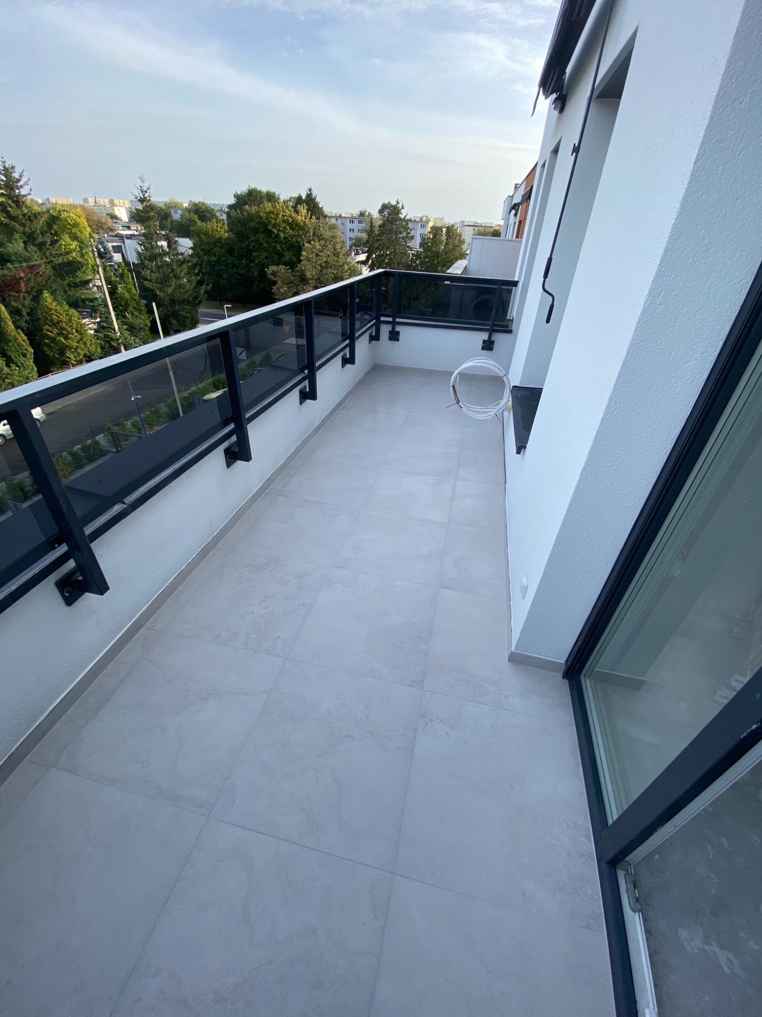 Jasne płytki na balkonie z czarną balustradą, widok na zieleń drzew i budynki w oddali, białe ściany budynku. Kabel elektryczny zwisający ze ściany.