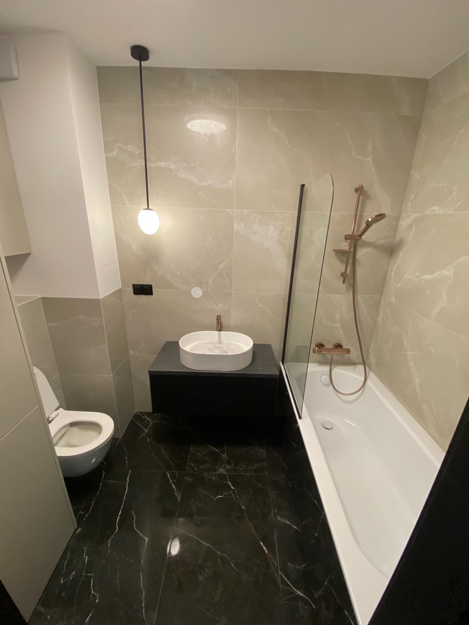 Nowoczesna łazienka z czarną marmurową podłogą, beżowymi płytkami imitującymi marmur na ścianach, białą umywalką nablatową na czarnej szafce, toaletą i białą wanną z parawanem oraz armaturą...