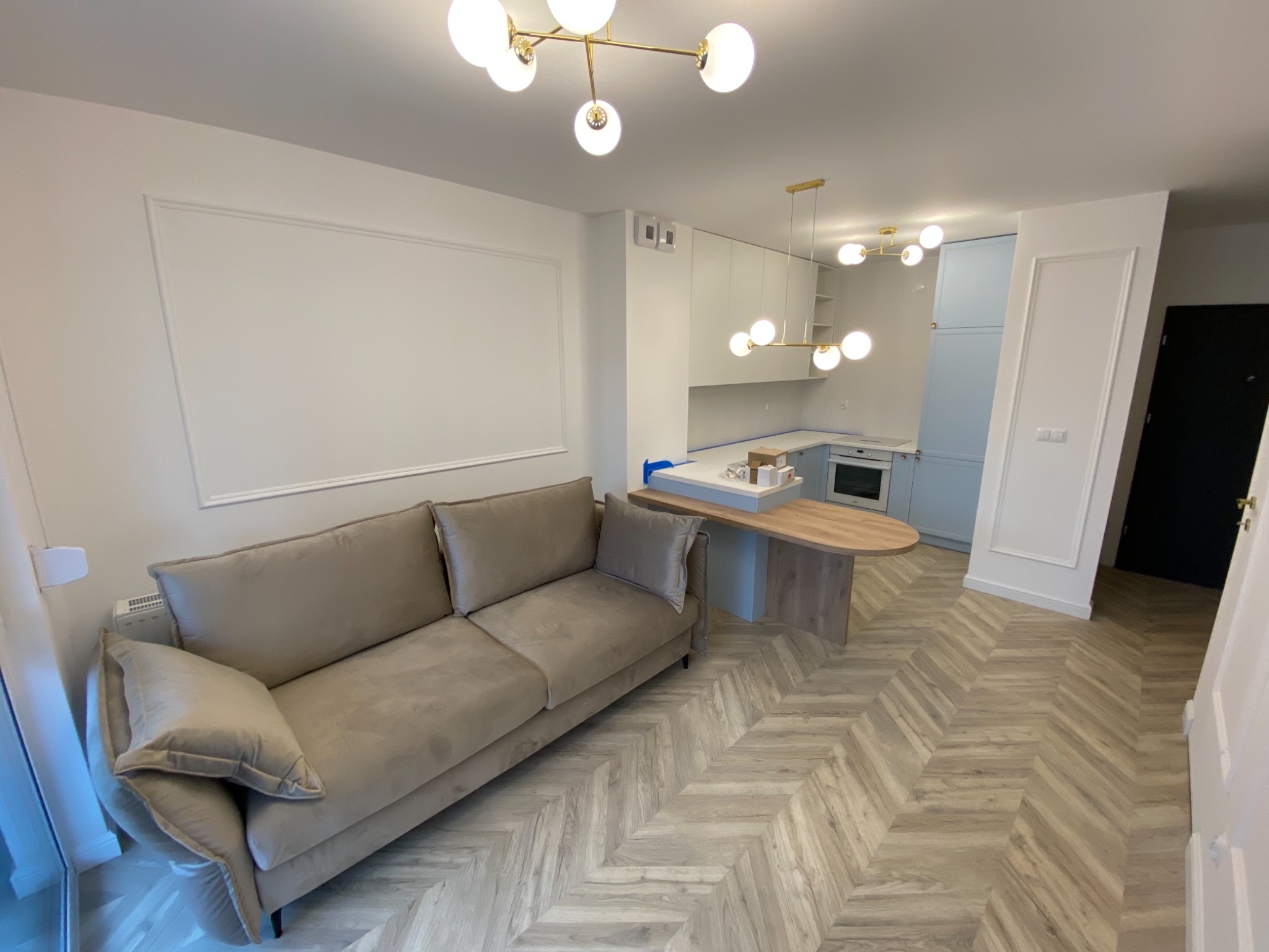 Salon z beżową sofą, panelami ułożonymi w jodełkę i nowoczesnym oświetleniem sufitowym w stylu glamour, widok na częściowo wykończoną kuchnię w tle.