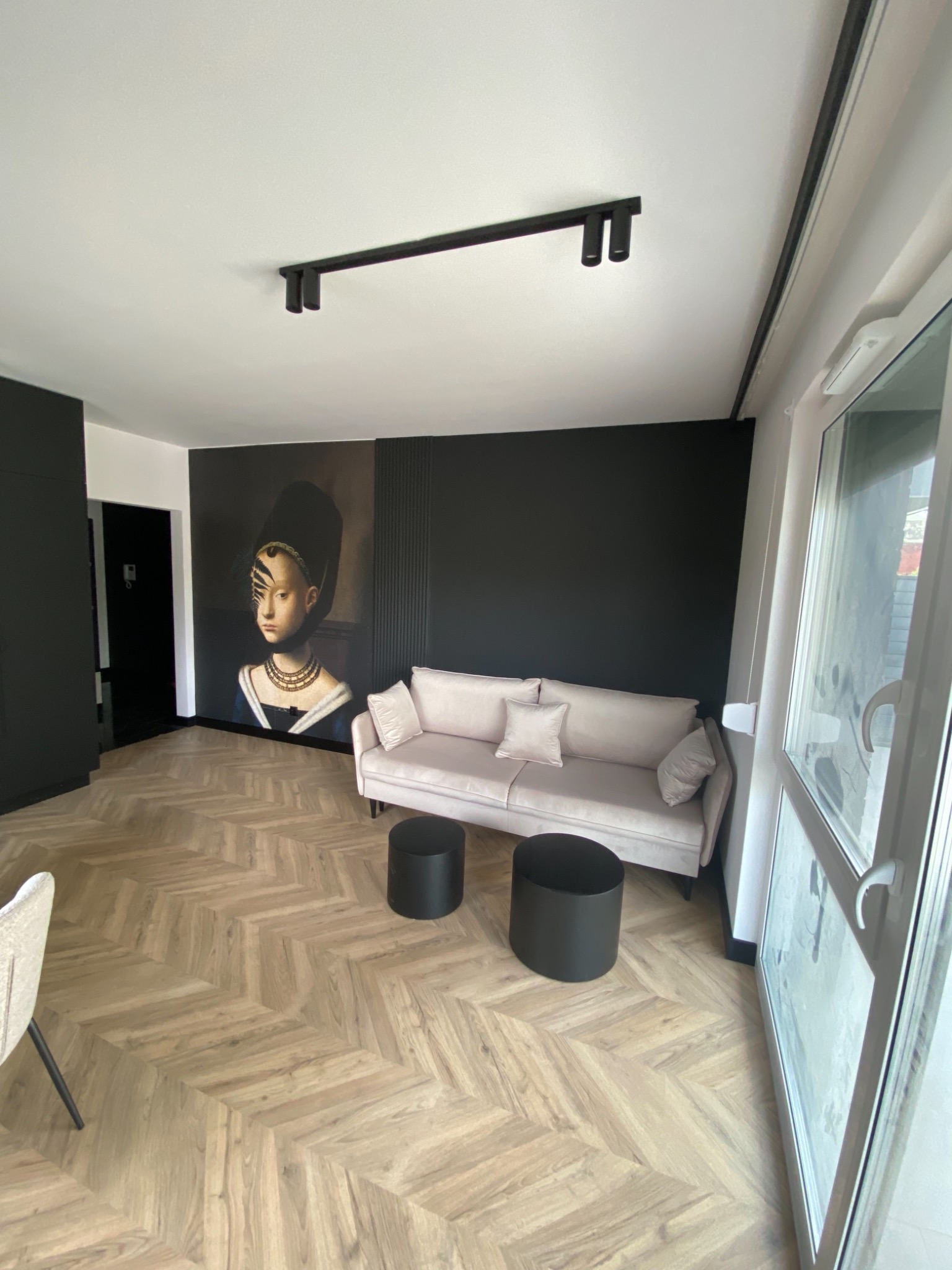 Jasny parkiet ułożony we wzór jodełki francuskiej w nowoczesnym salonie z czarną ścianą i portretem kobiety, beżowa sofa z poduszkami i czarne stołki.