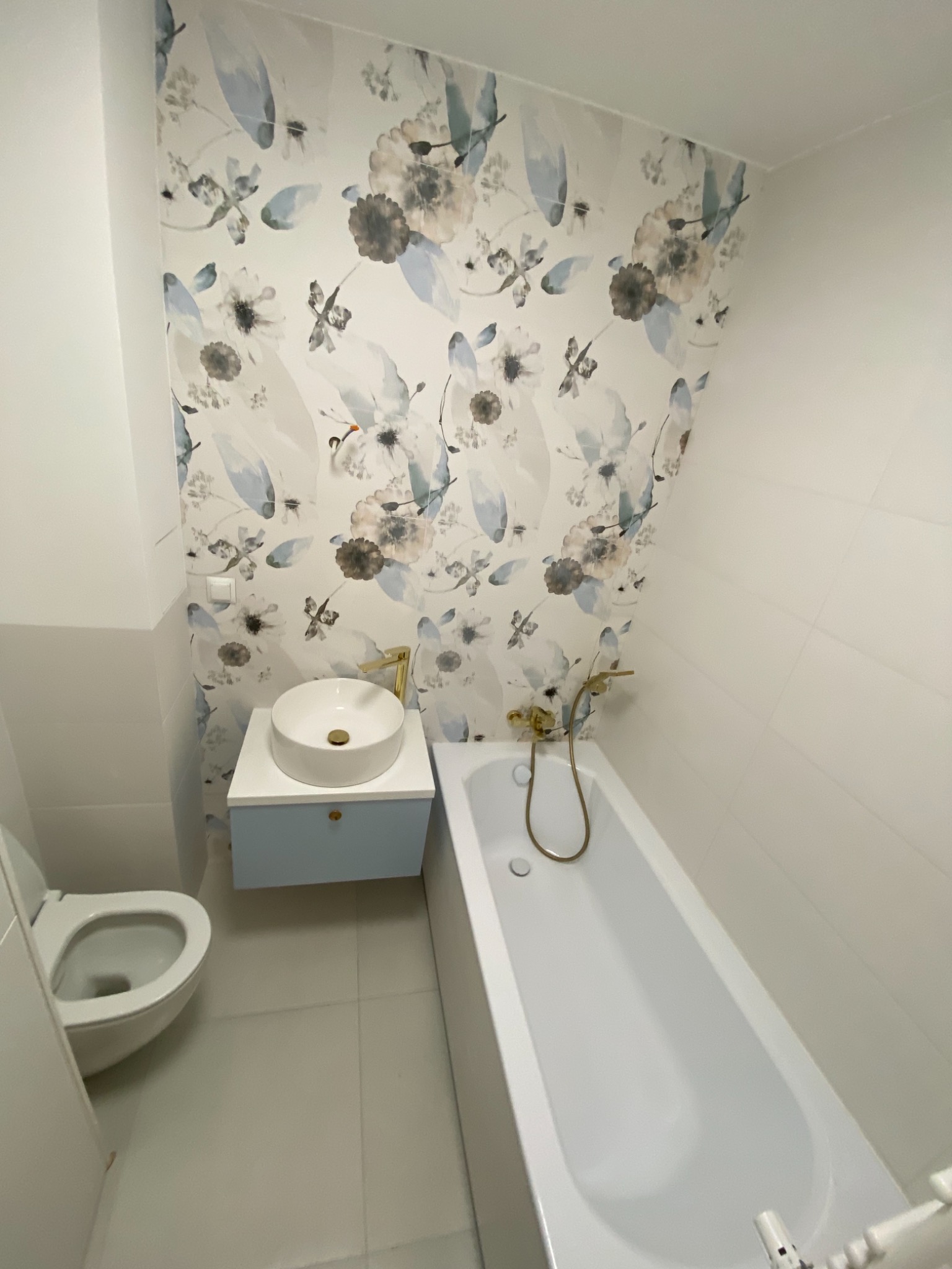 Wykończona łazienka z białą ceramiką, umywalką nablatową na błękitnej szafce, wanną i toaletą, akcentowana tapetą w kwiatowe wzory i złotą armaturą.