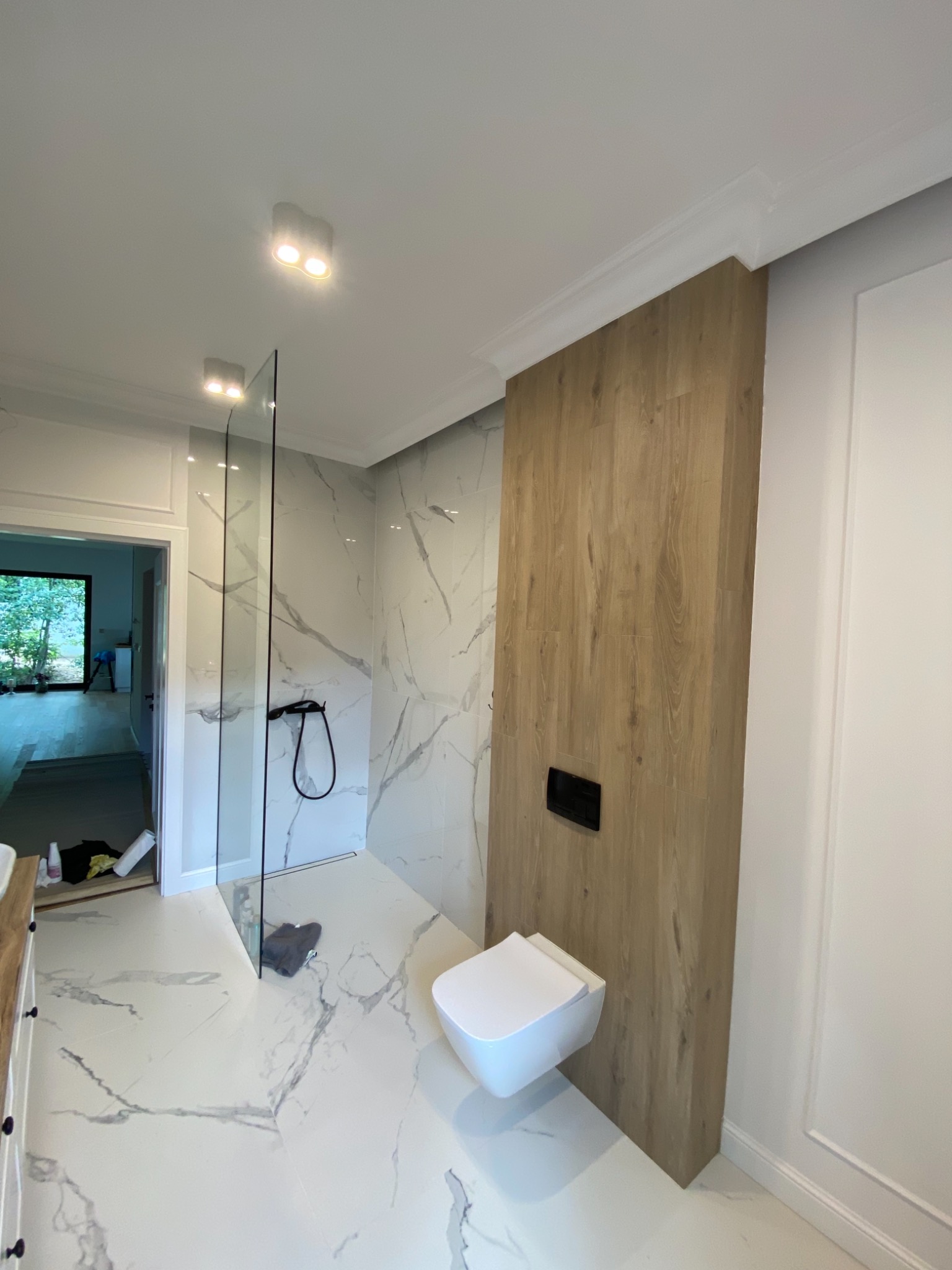 Nowoczesna łazienka z białą marmurową glazurą, kabiną prysznicową typu walk-in z czarną armaturą i podwieszaną toaletą obok drewnopodobnej obudowy stelaża.