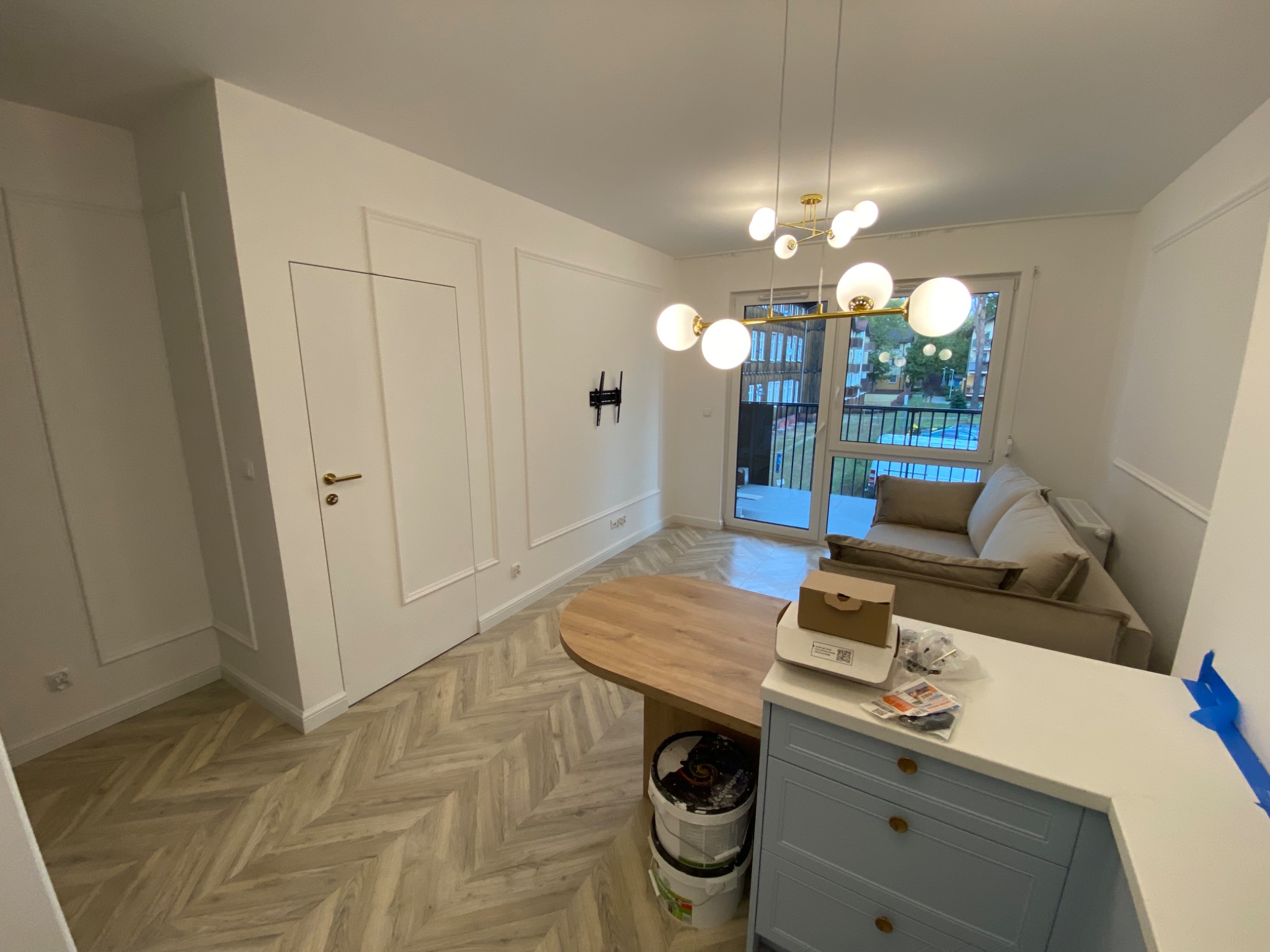 Jasne wnętrze mieszkania po remoncie: parkiet w jodełkę, białe ściany z dekoracyjnymi listwami, nowoczesna lampa wisząca, niebieska zabudowa kuchenna z akcesoriami na blacie i sofa w tle.