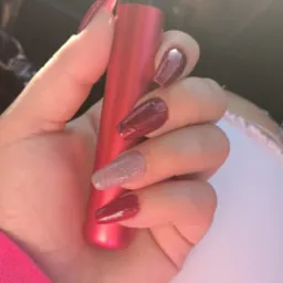 Dłoń z długimi, błyszczącymi paznokciami w odcieniach czerwieni i srebra, trzymająca czerwony e-papieros na tle siedzenia samochodowego.