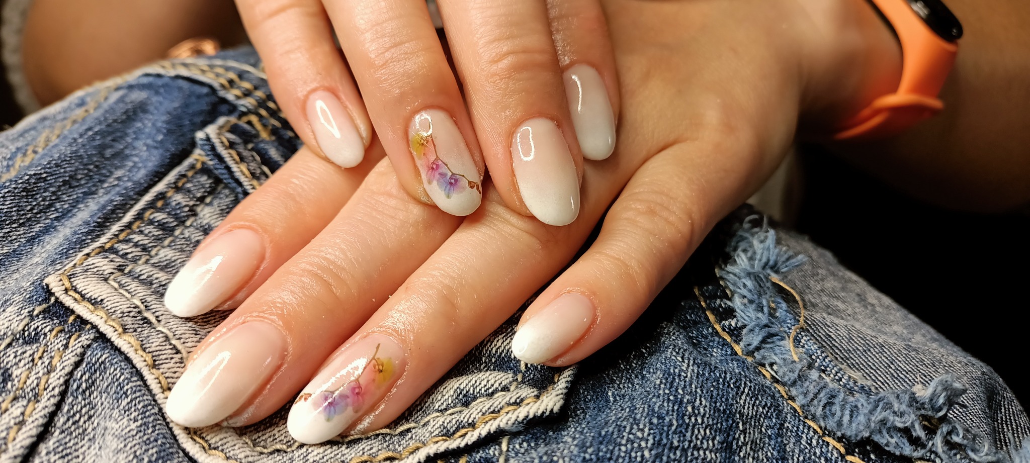 Delikatny manicure hybrydowy z efektem ombre i ręcznie malowanymi kwiatami, na tle dżinsowej kurtki.