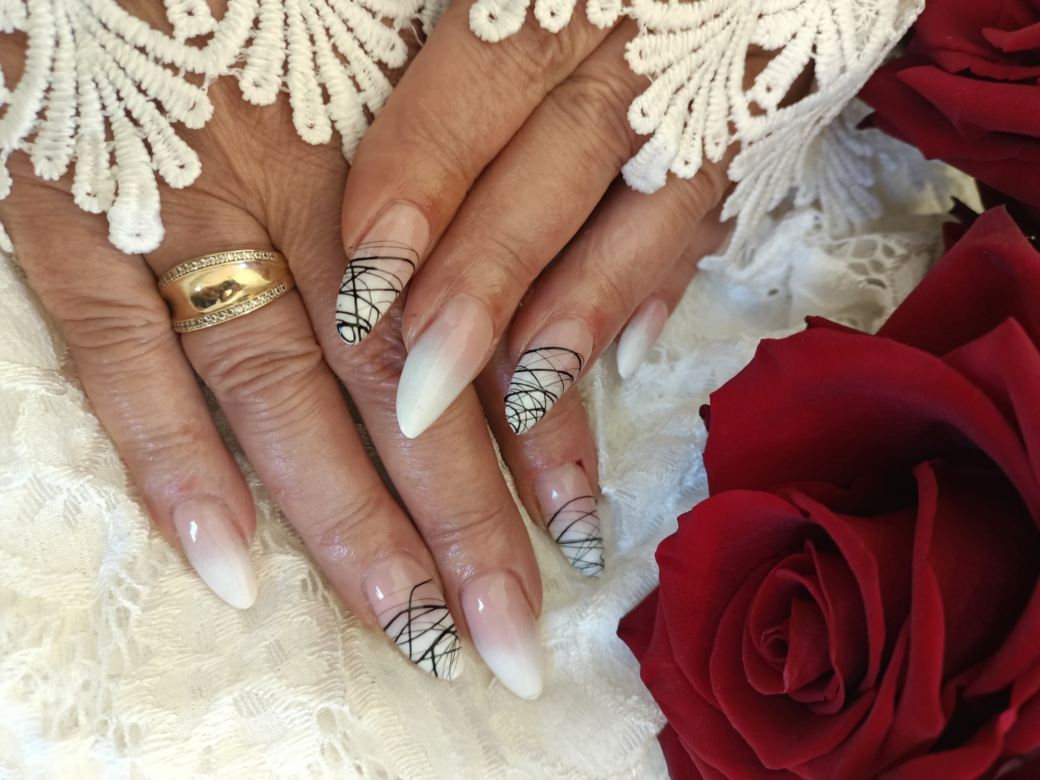 Dłonie z długimi, ombre paznokciami zdobionymi czarnymi, abstrakcyjnymi liniami na białym tle, złoty pierścionek na palcu, obok czerwona róża i biała koronka.
