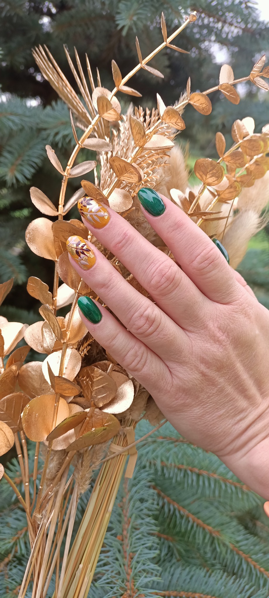 Dłoń z jesienną stylizacją paznokci: dwa paznokcie w kolorze zieleni butelkowej, pozostałe w odcieniach żółci z motywem liści i złotych płatków, na tle dekoracyjnego bukietu ze złotymi liśćmi...