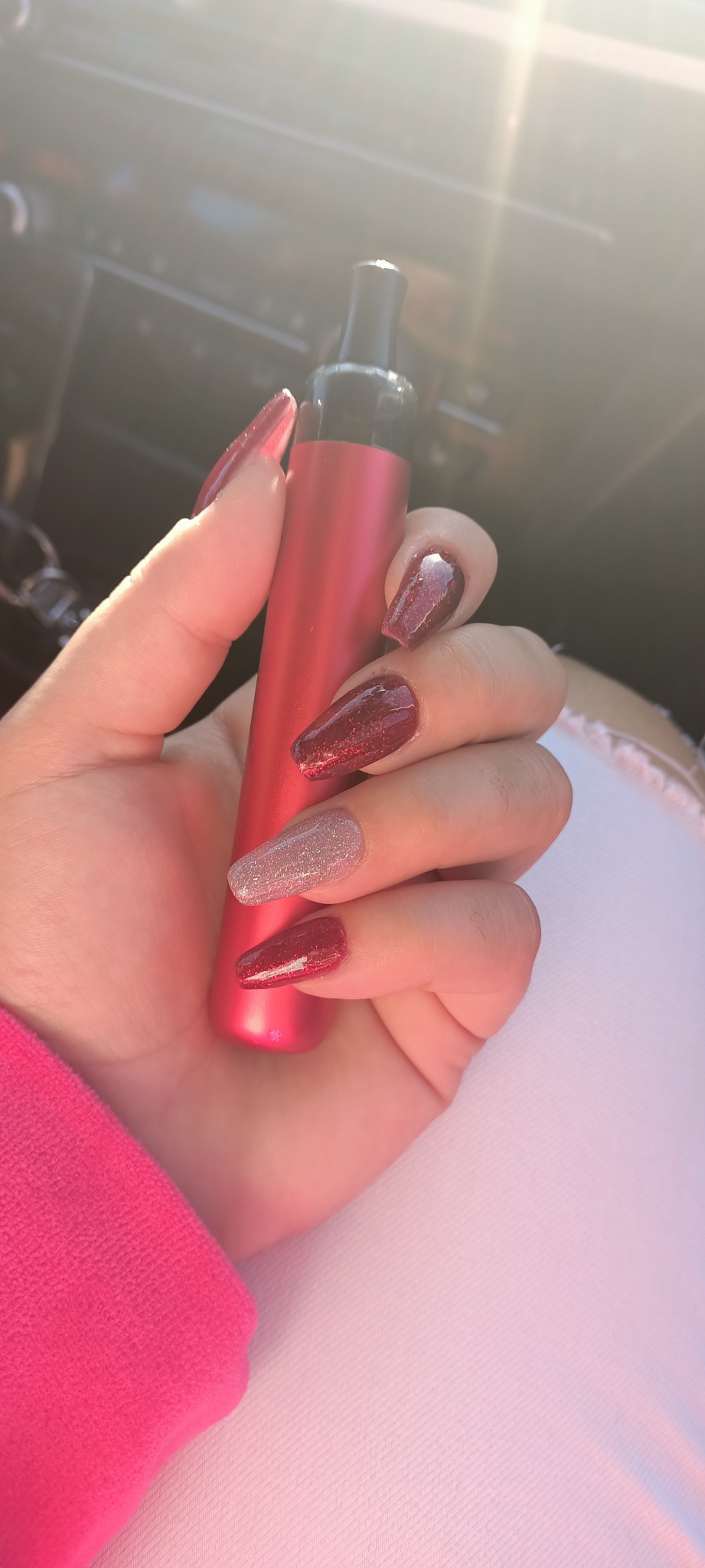 Dłoń z długimi, błyszczącymi paznokciami w odcieniach czerwieni i srebra, trzymająca czerwony e-papieros na tle siedzenia samochodowego.