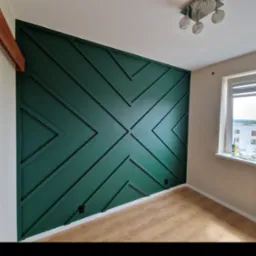 Ściana w pokoju z dekoracyjnym malowaniem w kolorze butelkowej zieleni, tworzącym geometryczny wzór z listew, drewniana podłoga, okno z widokiem na budynek mieszkalny.