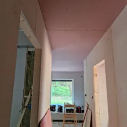 Korytarz w trakcie montażu płyt kartonowo-gipsowych, widoczna drabina, izolacja w ścianach, narzędzia i płyty czekające na montaż, sufit częściowo pokryty płytami w kolorze różowym.