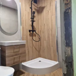 Nowoczesna łazienka z kabiną prysznicową typu walk-in, drewnopodobnymi płytkami na ścianach i podłodze, wiszącą szafką z umywalką oraz owalnym lustrem; widoczny fragment ściany w trakcie prac...