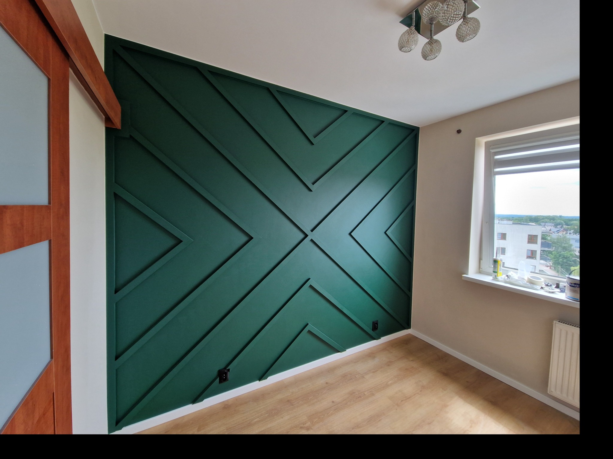 Ściana w pokoju z dekoracyjnym malowaniem w kolorze butelkowej zieleni, tworzącym geometryczny wzór z listew, drewniana podłoga, okno z widokiem na budynek mieszkalny.