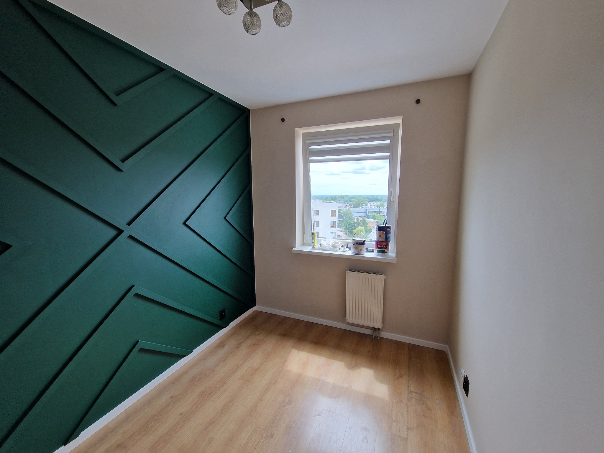 Pokój po remoncie: ściana z dekoracyjnymi, ciemnozielonymi listwami tworzącymi geometryczny wzór, jasne panele podłogowe, okno z widokiem na miasto, grzejnik pod oknem, sufit z nowoczesną lampą.