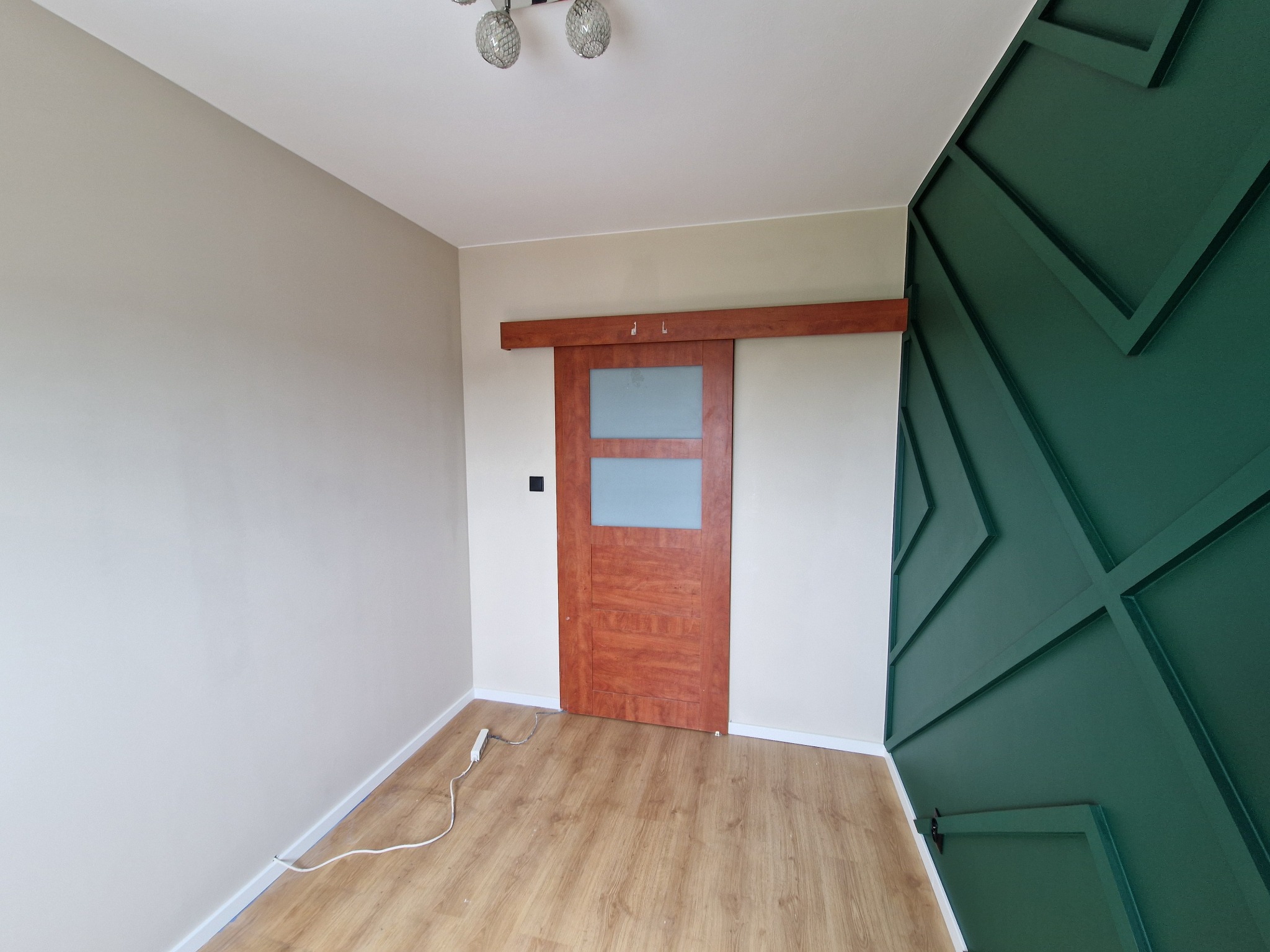 Wnętrze pokoju po remoncie: ściana pomalowana na beżowo, drewniane drzwi przesuwne z szybą, ściana z zielonymi, geometrycznymi panelami dekoracyjnymi, parkiet, widoczny kabel od listwy zasilającej.