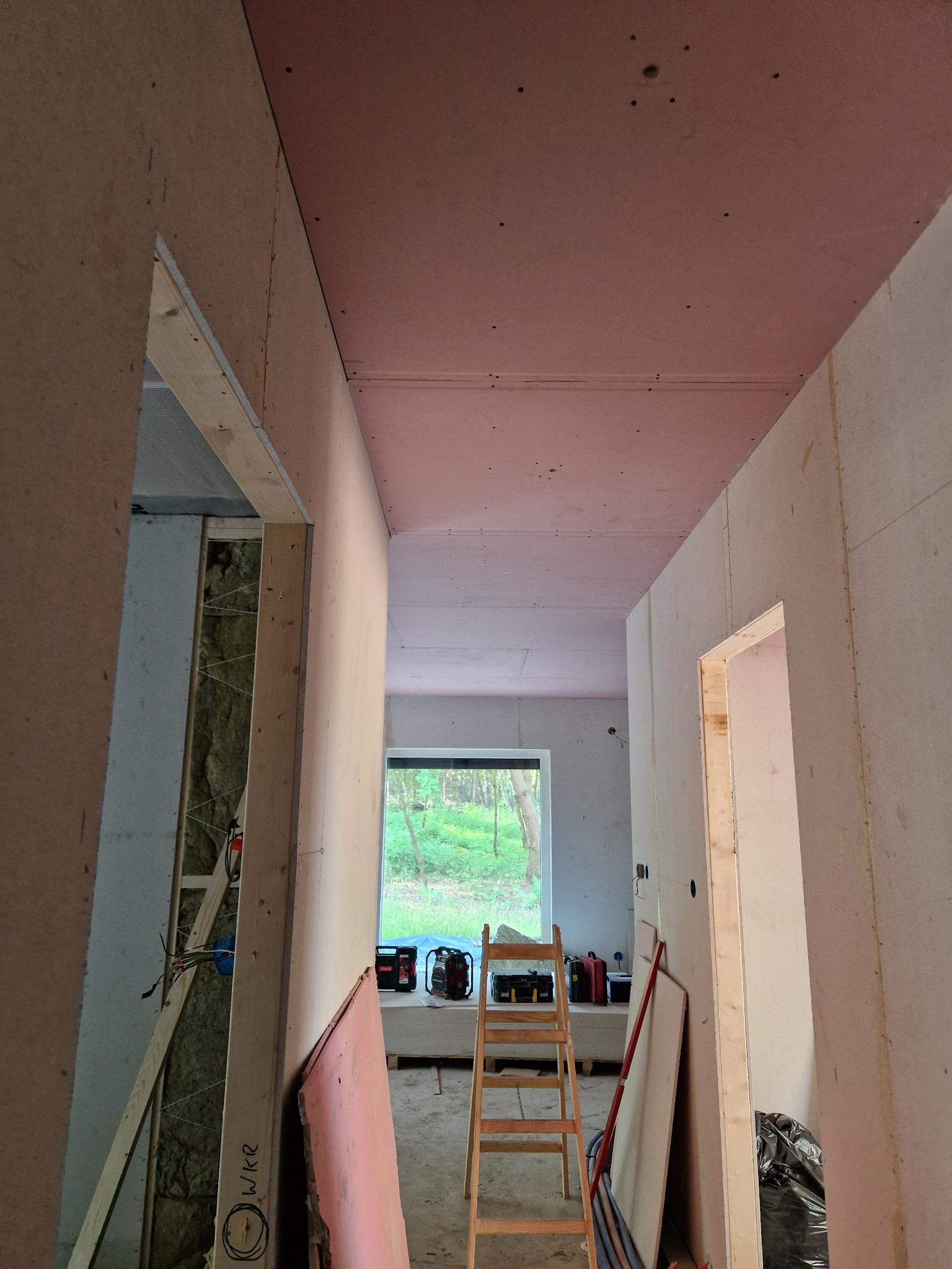 Korytarz w trakcie montażu płyt kartonowo-gipsowych, widoczna drabina, izolacja w ścianach, narzędzia i płyty czekające na montaż, sufit częściowo pokryty płytami w kolorze różowym.