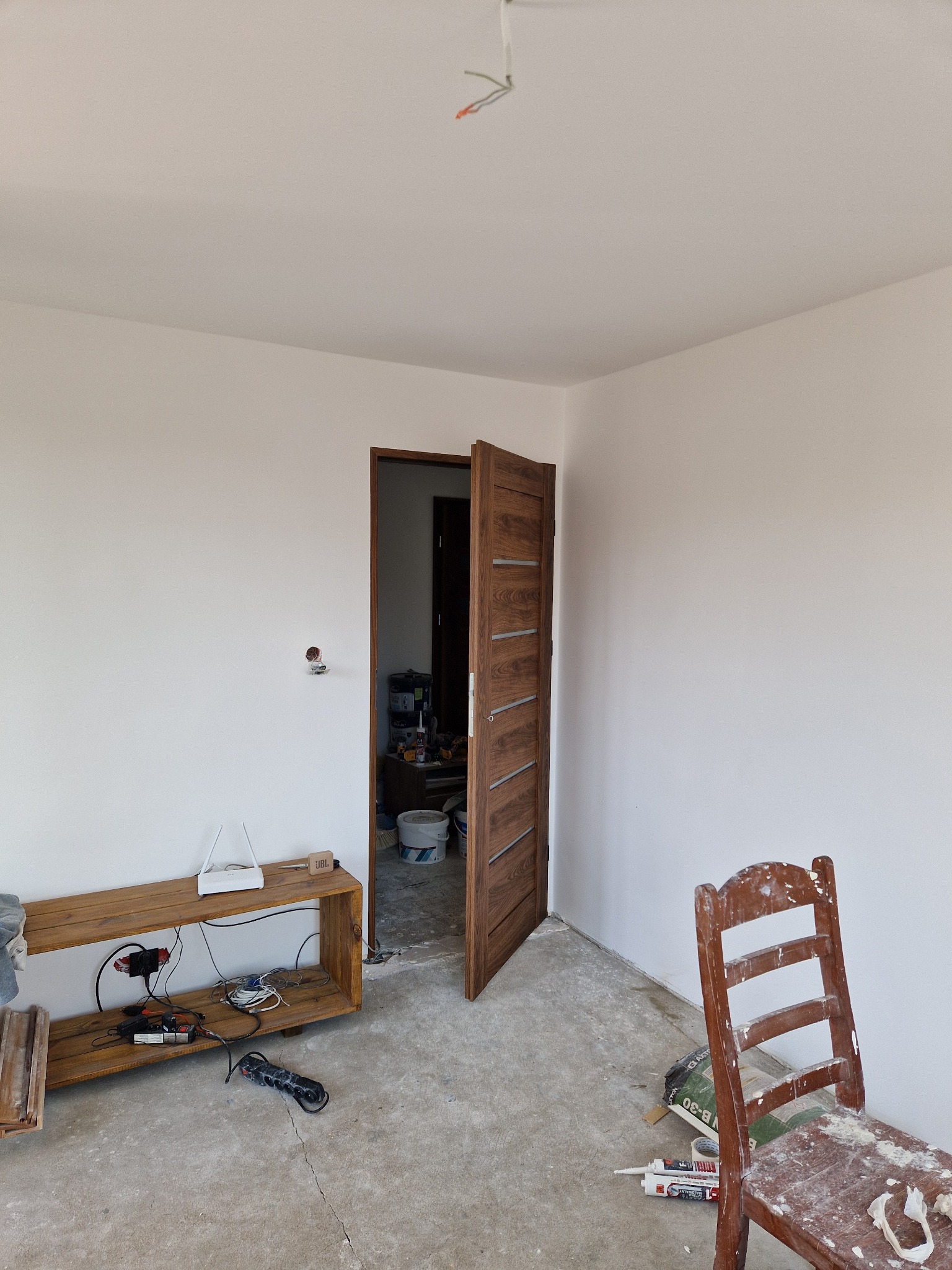 Pusta, pomalowana na biało ściana pokoju w trakcie remontu, widoczna drewniana półka z routerem i przedłużaczem, częściowo otwarte drzwi z poziomymi zdobieniami, krzesło robocze pobrudzone gipsem...