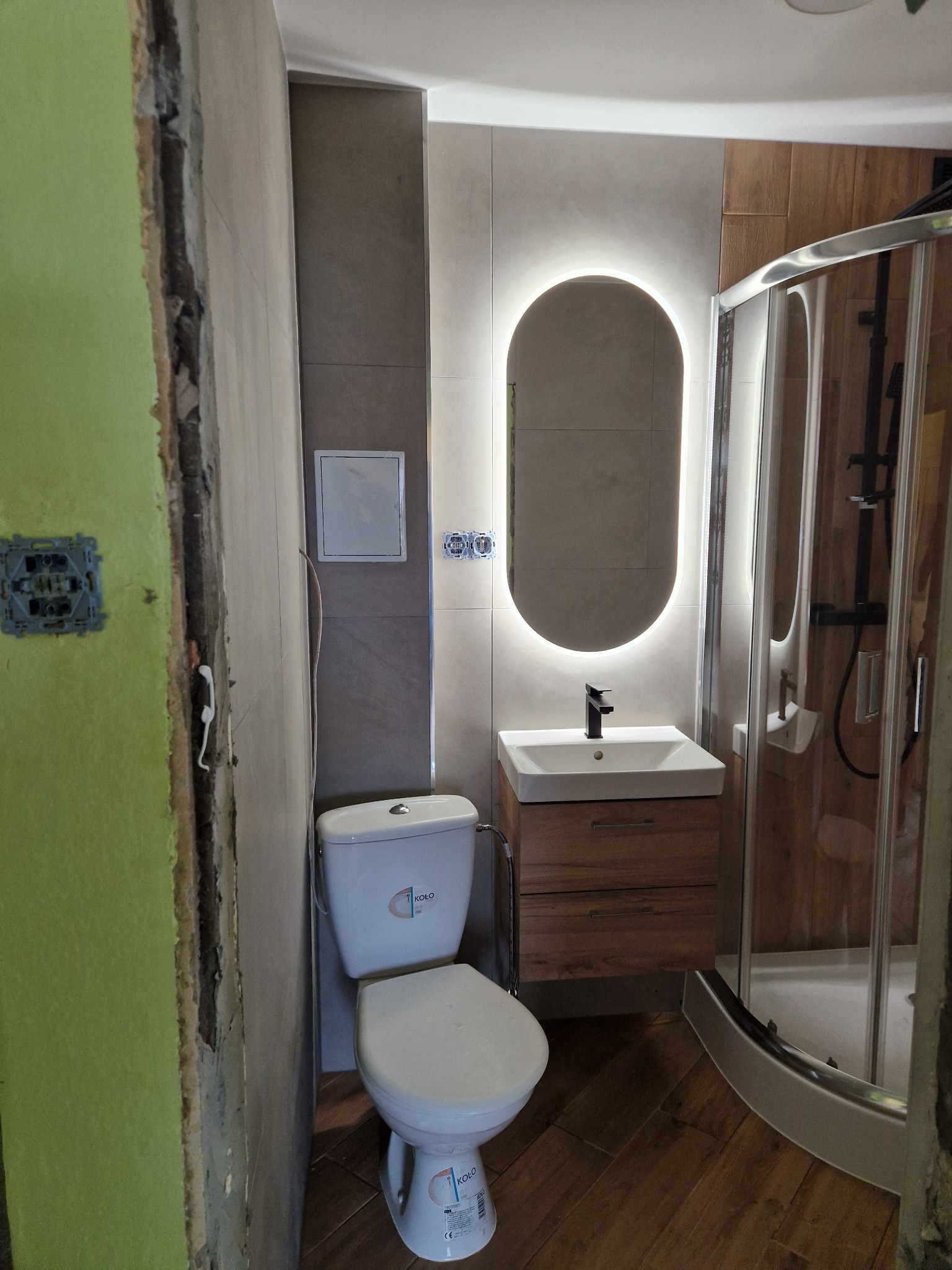 Wnętrze łazienki w trakcie remontu: widoczne surowe ściany z zieloną farbą obok nowej toalety KOLO, umywalki z szafką i kabiny prysznicowej z czarną armaturą, podświetlane owalne lustro.