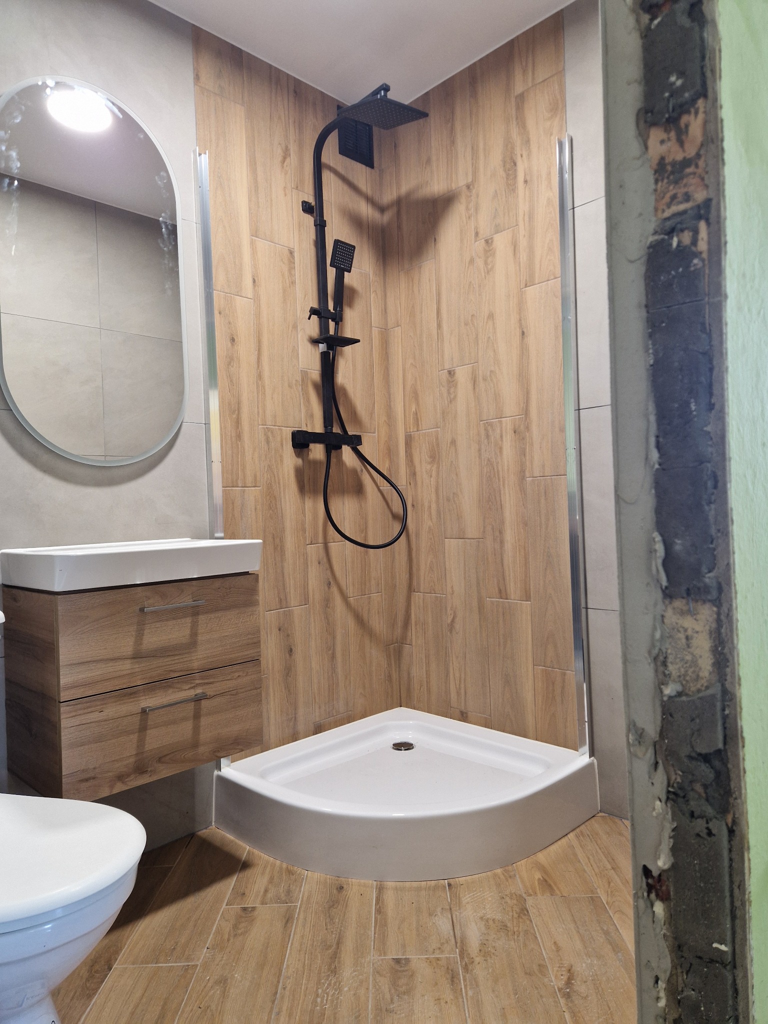 Nowoczesna łazienka z kabiną prysznicową typu walk-in, drewnopodobnymi płytkami na ścianach i podłodze, wiszącą szafką z umywalką oraz owalnym lustrem; widoczny fragment ściany w trakcie prac...