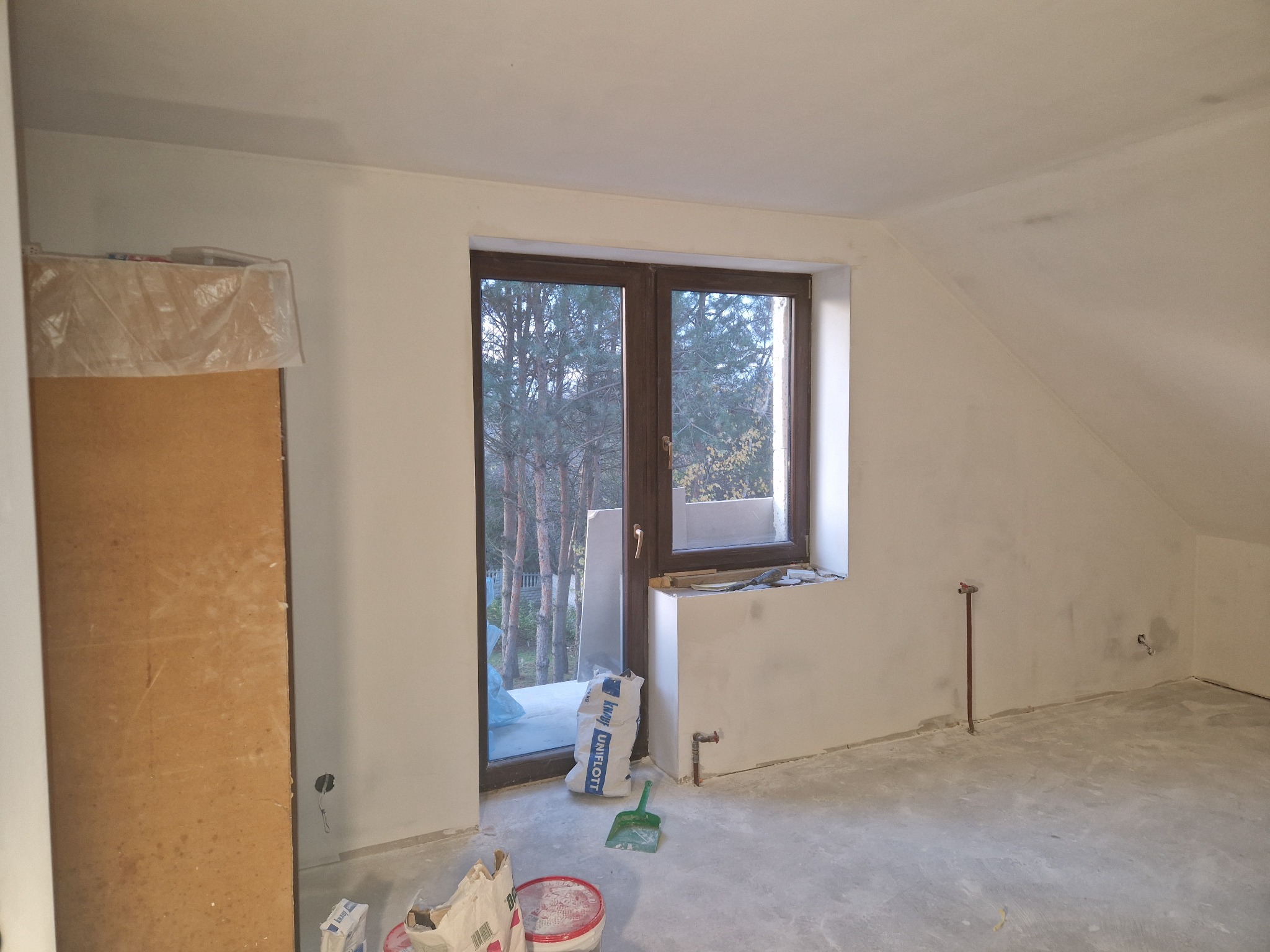 Poddasze w trakcie prac wykończeniowych: ściany po nałożeniu gładzi, widoczny worek z gładzią Knauf Uniflott, szpachelka, wiadro i wystające rury instalacji.