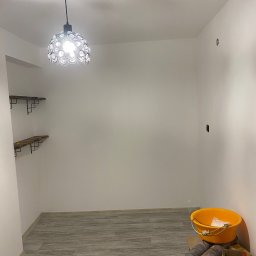 WRONA Usługi Remontowo Wykończeniowe Kacper Wrona - Pomalowane na biało puste pomieszczenie z ułożonymi panelami podłogowymi, dekoracyjną lampą sufitową i dwiema drewnianymi półkami na ścianie. W rogu stoi żółte wiadro, wałki i inne akcesoria...
