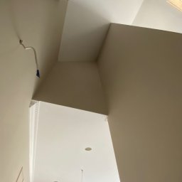 WRONA Usługi Remontowo Wykończeniowe Kacper Wrona - Pomieszczenie po malowaniu, jasne ściany i sufit, widoczne elementy dekoracyjne i fragment drabiny malarskiej. Świeżo pomalowane wnętrze z detalami wykończeniowymi.