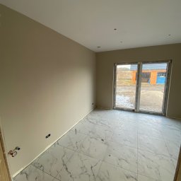 WRONA Usługi Remontowo Wykończeniowe Kacper Wrona - Jasne wnętrze po remoncie: beżowe ściany, białe marmurowe płytki podłogowe, duże okno z widokiem na zewnątrz. Widoczne gniazdka elektryczne i instalacja wodna.