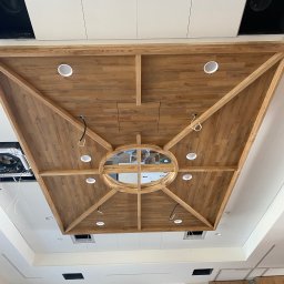 WRONA Usługi Remontowo Wykończeniowe Kacper Wrona - Dekoracyjny sufit podwieszany z drewnianymi panelami i okrągłym otworem w centrum, oświetlony punktowymi lampami, widok z dołu.