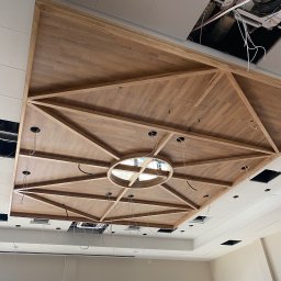 WRONA Usługi Remontowo Wykończeniowe Kacper Wrona - Dekoracyjny, drewniany sufit podwieszany z geometrycznym wzorem i okrągłym elementem centralnym. Widoczne otwory na oświetlenie i fragmenty instalacji.