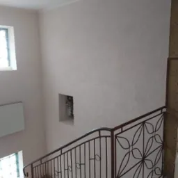 Klatka schodowa po malowaniu, widoczne świeżo pomalowane ściany w jasnym odcieniu, dekoracyjna metalowa balustrada z motywem kwiatowym, okno z luksferami i wnęka z licznikiem.