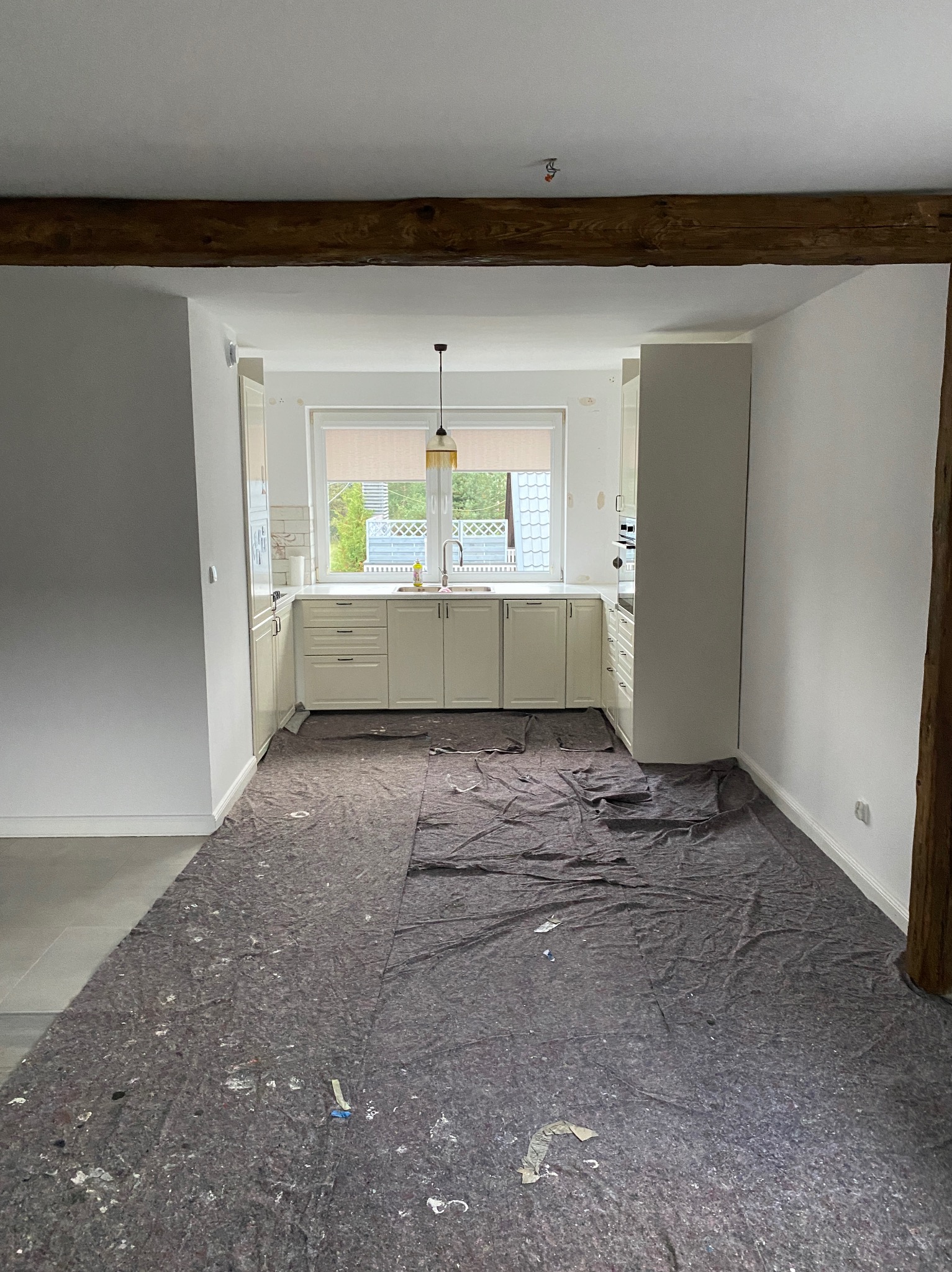 Wnętrze kuchni w trakcie prac wykończeniowych, podłoga zabezpieczona szarą matą, widoczne drewniane belki stropowe i białe ściany po malowaniu.
