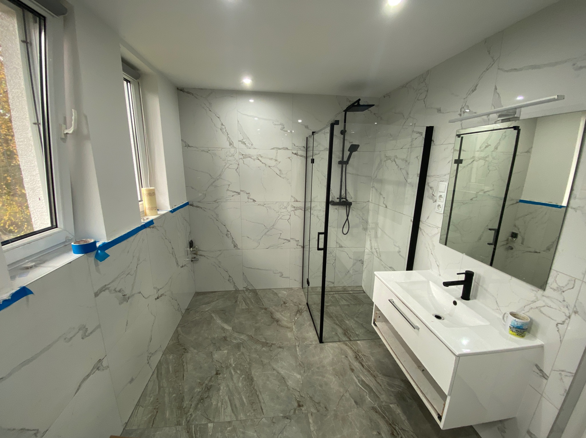 Nowoczesna łazienka po remoncie: białe płytki imitujące marmur, szara podłoga, czarna kabina prysznicowa i biała szafka z umywalką. Widoczne zabezpieczenia okna.