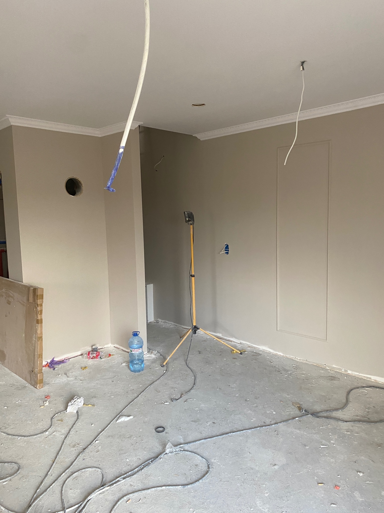 Wnętrze w trakcie prac wykończeniowych: pomalowane ściany w beżowym odcieniu, widoczne kable, lampa na statywie i otwór w ścianie. Podłoga surowa, w trakcie przygotowań.